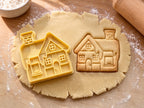 Butter-gelber Haus-Ausstecher mit Prägung neben gebackenem Keks mit Schneedach, Ziegelkamin und Sprossenfenstern auf hellem Holz