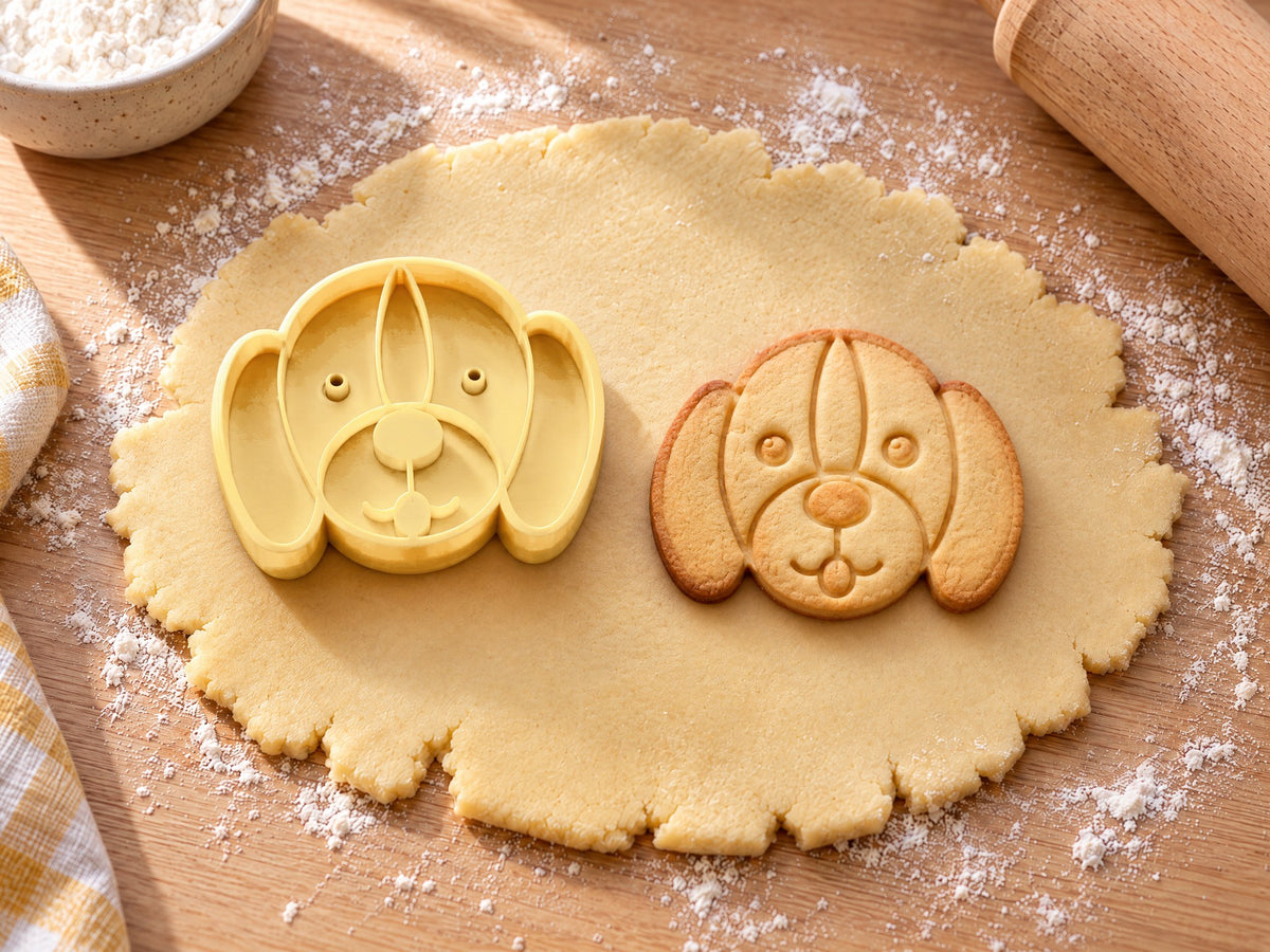 Butter-gelber Ausstecher in Form eines Hundekopfs neben gebackenem Hundekeks mit geprägtem Gesicht und Schlappohren auf ausgerolltem Teig und hellem Holz