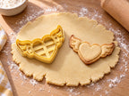 Butter-gelber Herz-Ausstecher mit Prägung neben gebackenem Keks: Herz mit Innenlinie und zwei gefiederten Flügeln auf hellem Holz