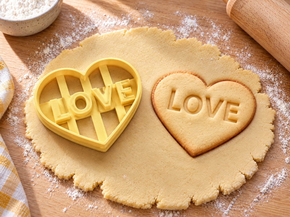 Butter-gelber Herz-Ausstecher mit Prägung neben gebackenem Keks in Herzform mit eingeprägtem Schriftzug LOVE auf hellem Holz