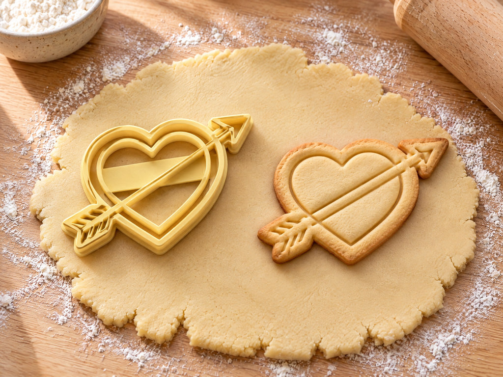 Butter-gelber Herz-Ausstecher mit Prägung neben gebackenem Keks: Herz mit Innenlinie, schräg von einem Pfeil durchbohrt, auf hellem Holz