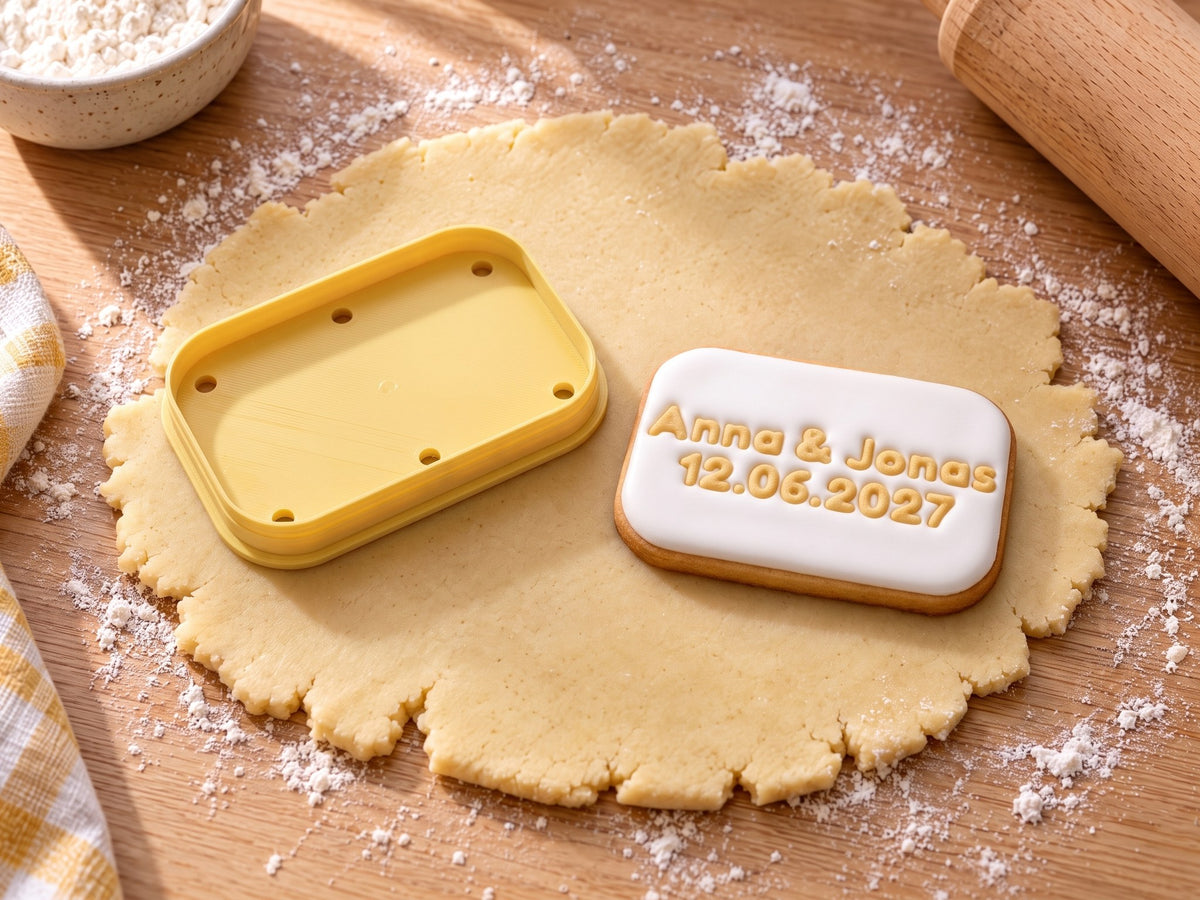 Butter-gelber Plaketten-Ausstecher neben gebackener Keks-Plakette mit weißem Zuckerguss und geprägtem Text „Anna & Jonas 12.06.2027“ auf ausgerolltem Teig und hellem Holz