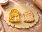 Butter-gelber Ausstecher mit Prägung: sitzender Welpe mit Weihnachtsmütze, daneben Keks mit Schneeflockenbommel auf hellem Holz