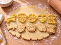 Set Hunde-Liebe: 5 butter-gelbe Ausstecher mit Prägung nebeneinander mit gebackenen, geprägten Keksen auf ausgerolltem Teig und hellem Holz