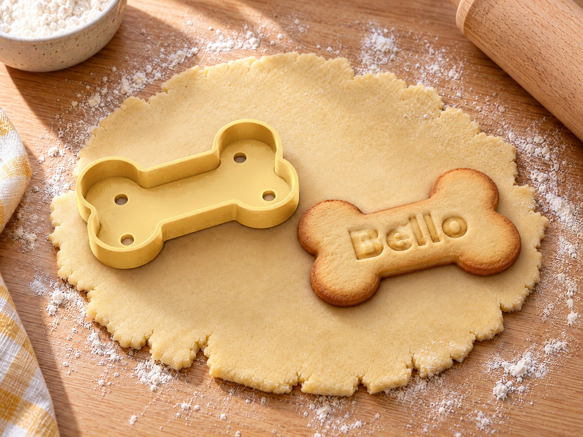 Butter-gelber Hundeknochen-Ausstecher neben gebackenem Knochenkeks mit geprägtem Namen „Bello“ auf ausgerolltem Teig und hellem Holz