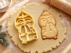 Butter-gelber Pinguin-Ausstecher mit Prägung neben gebackenem Keks: kichernder Pinguin mit Bommelmütze und Schal auf hellem Holz