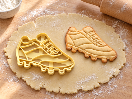 Fußballschuh-Ausstecher mit Prägung neben gebackenem Keks mit Schnürung, Seitenstreifen und Stollen auf hellem Holz