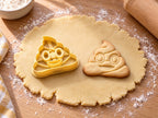 Butter-gelber Kackhaufen-Emoji-Ausstecher mit Prägung neben gebackenem Keks mit Windungen, Kulleraugen und Grinsen auf hellem Holz