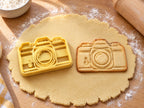 Butter-gelber Kamera-Ausstecher mit Prägung neben gebackenem Keks mit Objektivringen, Sucher und Auslöser auf hellem Holz
