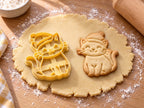 Butter-gelber Katzen-Ausstecher mit Prägung neben gebackenem Keks: Katze mit Bommelmütze und Schnurrhaaren auf hellem Holz