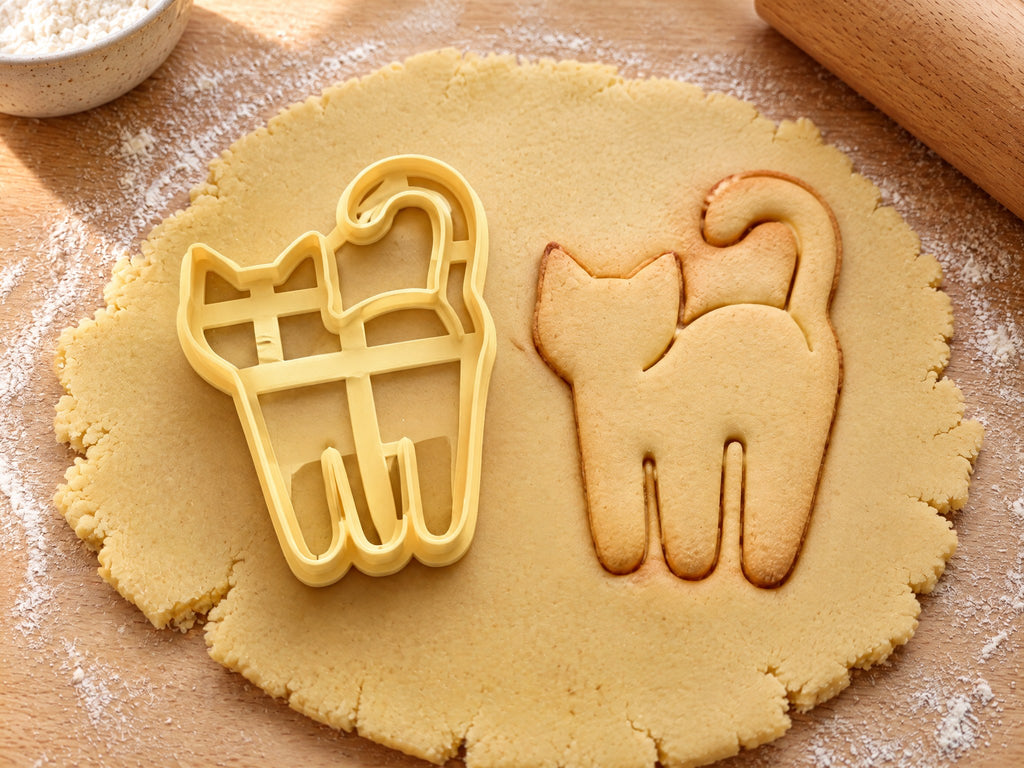 Butter-gelber Katzen-Ausstecher neben gebackenem Keks in Form einer stehenden Katze mit hochgebogenem Schwanz auf hellem Holz