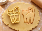 Butter-gelber Katzen-Ausstecher neben gebackenem Keks in Form einer stehenden Katze mit hochgebogenem Schwanz auf hellem Holz