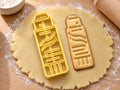 Butter-gelber Klebestift-Ausstecher mit Prägung neben gebackenem Keks mit Kappe, Wellenetikett und Drehrad auf hellem Holz