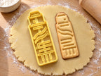 Butter-gelber Klebestift-Ausstecher mit Prägung neben gebackenem Keks mit Kappe, Wellenetikett und Drehrad auf hellem Holz