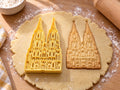 Butter-gelber Kölner-Dom-Ausstecher mit Prägung neben gebackenem Keks mit zwei Türmen und Spitzbogenfenstern auf hellem Holz