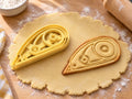 Butter-gelber Korrekturroller-Ausstecher mit Prägung neben gebackenem Keks mit zwei Spulen und schmaler Spitze auf hellem Holz