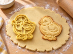 Butter-gelber Küken-Ausstecher mit Prägung neben gebackenem Keks mit flauschigem Gefieder, Flügel und Füßchen auf hellem Holz