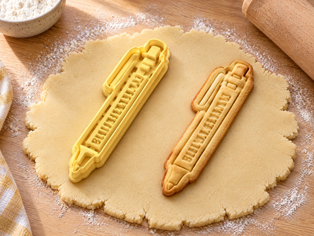 Butter-gelber Kugelschreiber-Ausstecher mit Prägung neben schmalem gebackenem Keks mit Clip und Druckknopf auf hellem Holz