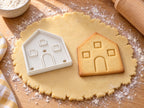 Giebelwand-Ausstecher mit Prägung neben gebackenem Lebkuchen-Keks mit Bogentür und Fenstern auf hellem Holz