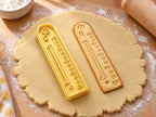 Butter-gelber Lineal-Ausstecher mit Prägung neben gebackenem Keks mit Skala und Aufhängeloch auf hellem Holz