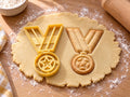 Butter-gelber Medaillen-Ausstecher mit Prägung neben gebackenem Keks mit V-Band, Öse und Stern auf hellem Holz