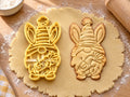 Butter-gelber Osterwichtel-Ausstecher mit Prägung neben Keks mit gestreifter Mütze, Hasenohren und Karotte auf hellem Holz