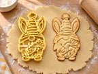 Butter-gelber Osterwichtel-Ausstecher mit Prägung neben Keks mit gestreifter Mütze, Hasenohren und Karotte auf hellem Holz