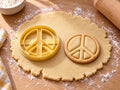 Butter-gelber Peace-Zeichen-Ausstecher mit Prägung neben gebackenem Keks mit breiten Balken und schmalem Ring auf hellem Holz