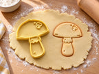 Butter-gelber Pilz-Ausstecher mit Prägung neben gebackenem Keks mit Manschette am Stiel und glattem Hut auf hellem Holz