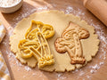 Butter-gelber Ausstecher mit Prägung: zwei Pilze im Grasbüschel, daneben gebackener Keks mit gesprenkelten Hüten auf hellem Holz