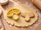 Butter-gelber Pilz-Ausstecher mit Prägung neben gebackenem Keks mit Strahlenlamellen und dickem Stiel auf hellem Holz
