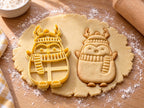 Butter-gelber Pinguin-Ausstecher mit Prägung neben gebackenem Keks mit Bommelmütze, Rentiergeweih und Streifenschal auf hellem Holz
