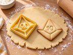 Butter-gelber Ausstecher mit Prägung als Zehnseiter-Würfel neben gebackenem Keks mit Kantenlinien und der Zahl 10 auf hellem Holz