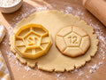 Butter-gelber Ausstecher mit Prägung als Zwölfseiter-Würfel neben gebackenem Keks mit Fünfeck und Zahl 12 auf hellem Holz