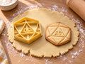 Butter-gelber Ausstecher mit Prägung als Zwanzigseiter-Würfel neben gebackenem Keks mit Dreiecksfacetten und der Zahl 20 auf hellem Holz