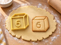 Butter-gelber Würfel-Ausstecher mit Prägung neben gebackenem Keks in Würfelform mit drei Seitenflächen und großer 6 auf hellem Holz