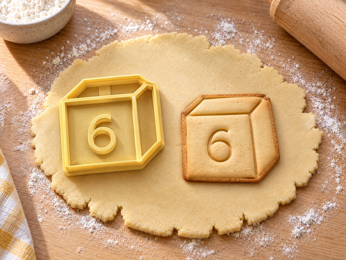 Butter-gelber Würfel-Ausstecher mit Prägung neben gebackenem Keks in Würfelform mit drei Seitenflächen und großer 6 auf hellem Holz