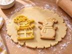 Butter-gelber Schneemann-Ausstecher mit Prägung neben gebackenem Keks mit langer Karottennase, Pudelmütze und Zweigarmen auf hellem Holz