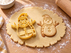 Butter-gelber Schneemann-Ausstecher mit Prägung neben gebackenem Keks: winkender Schneemann mit Bommelmütze und Schal auf hellem Holz