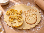 Butter-gelber Schneemann-Ausstecher mit Prägung neben gebackenem Keks mit Ringelmütze, Fransenschal und Zuckerstange auf hellem Holz