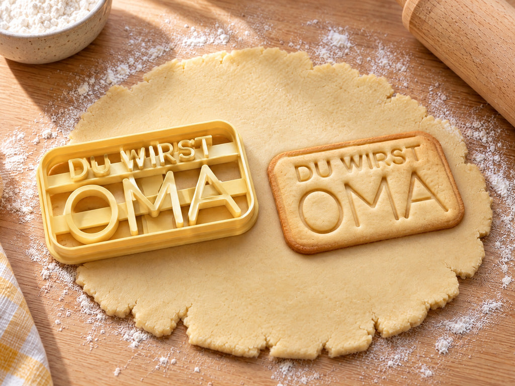 Butter-gelber Ausstecher mit Prägung neben rechteckigem Keks mit Schriftzug „DU WIRST OMA“ im Rahmen auf hellem Holz