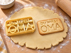 Butter-gelber Ausstecher mit Prägung neben rechteckigem Keks mit Schriftzug „DU WIRST OMA“ im Rahmen auf hellem Holz