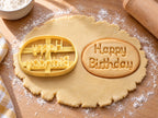 Butter-gelber Ausstecher mit Prägung „Happy Birthday“ neben ovalem Keks mit zweizeiligem Schriftzug auf hellem Holz