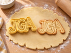 Butter-gelber JGA-Ausstecher mit Prägung neben Keks mit hohen Serifenbuchstaben JGA in doppelter Kontur auf hellem Holz