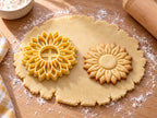 Butter-gelber Sonnenblumen-Ausstecher mit Prägung neben gebackenem Keks mit doppeltem Blütenkranz und glatter Mitte auf hellem Holz