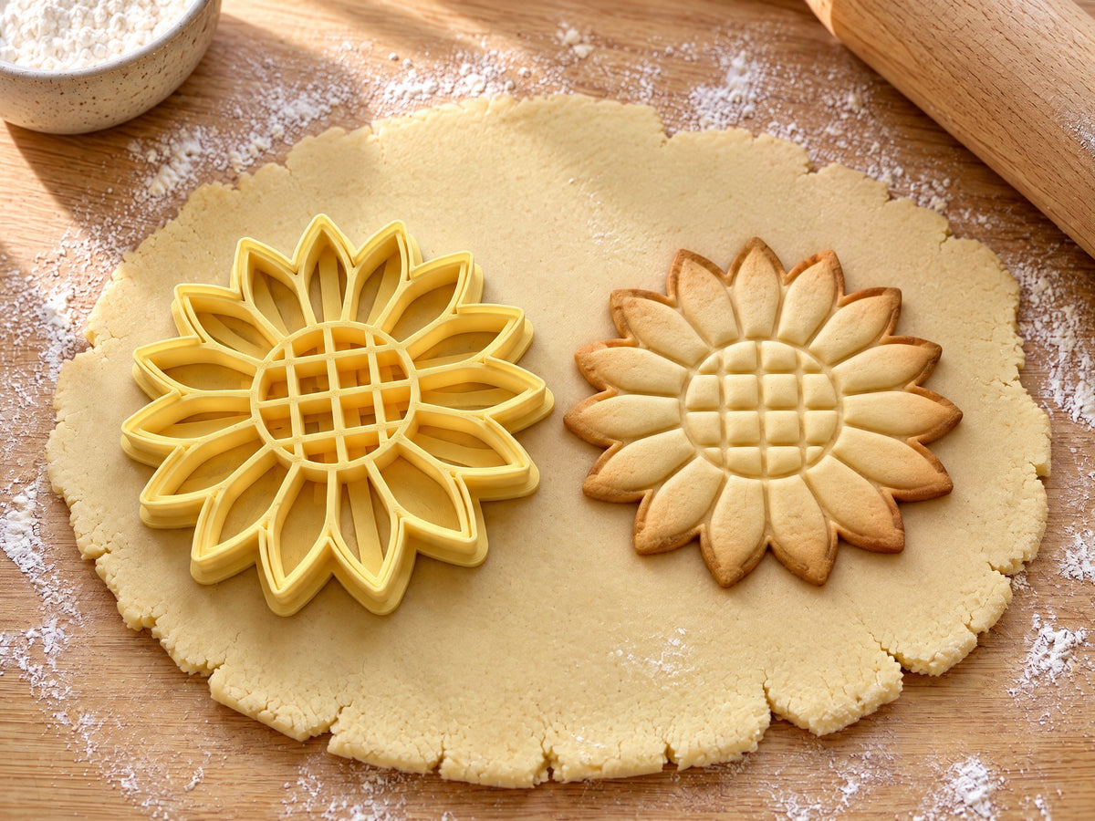 Butter-gelber Sonnenblumen-Ausstecher mit Prägung neben gebackenem Keks mit Blütenkranz und gegitterter Kernmitte auf hellem Holz