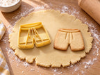 Butter-gelber Sporthose-Ausstecher mit Prägung neben gebackenem Keks mit Bund, Kordel und zwei Hosenbeinen auf hellem Holz