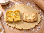 Butter-gelber Tassenrand-Ausstecher mit Prägung in Buchform neben gebackenem Keks mit Textzeilen und Herzen auf hellem Holz