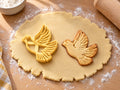 Butter-gelber Tauben-Ausstecher mit Prägung neben gebackenem Keks mit fliegender Taube und gefächerten Flügelfedern auf hellem Holz