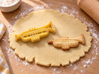 Butter-gelber Trompeten-Ausstecher mit Prägung neben gebackenem Keks mit drei Ventilen und geprägter Rohrschleife auf hellem Holz